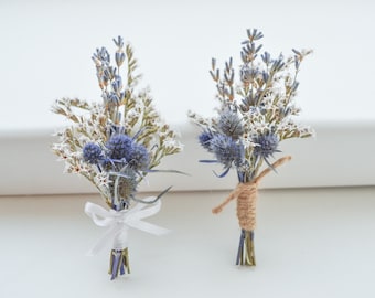 May include: Two small, dried flower bouquets. Each bouquet features blue thistle, lavender, and white star-shaped flowers. One is tied with a white ribbon, the other with twine. Perfect for weddings or special events.