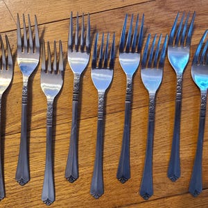 May include: A collection of nine silver forks with ornate handle detailing, arranged on a wooden surface. The forks have tines and decorative patterns on the handles. The forks are a vintage style, suitable for a formal dining setting.