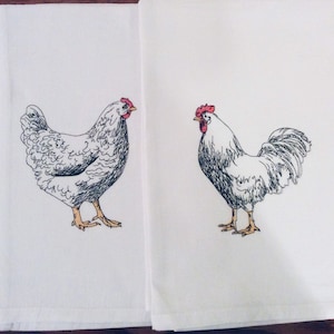May include: Two white kitchen towels with embroidered chicken and rooster designs. The chicken is on the left, and the rooster is on the right. Both have red combs and yellow feet. The towels are folded and placed side by side.