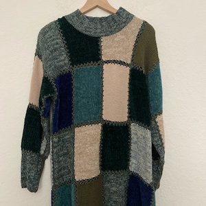 May include: A patchwork sweater in shades of green, blue, beige, and olive green hangs on a wooden hanger. The sweater features a crew neck and long sleeves. The design is made up of various rectangular patches sewn together with a contrasting thread.