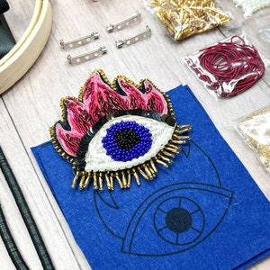 Könnte beinhalten: Ein Bastelset zum Herstellen einer bösen Augenbrosche. Das Set enthält eine blaue Filzbasis, ein böses Auge aus Perlen, rosa und goldene Perlen sowie eine Vielzahl weiterer Materialien.
