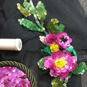 Puede incluir: Primer plano de un diseño de bordado floral con lentejuelas rosas, amarillas y verdes. El diseño está sobre un fondo de tela negra.