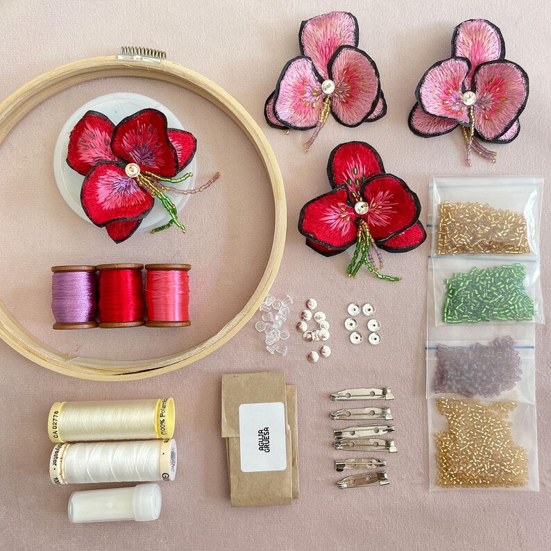 May include: A craft kit for making embroidered flower brooches. The kit includes pink and red embroidered flowers, beads, thread, needles, and pins. The flowers are arranged on a wooden embroidery hoop.