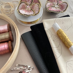 May include: A craft supply collection featuring two embroidered pink flower brooches with gold beaded edges, spools of pink and metallic thread, a wooden embroidery hoop, gold wire, black leather and velvet fabric swatches, and silver safety pins.