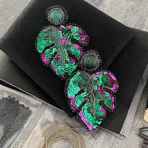 May include: A pair of handcrafted earrings featuring embroidered monstera leaf shapes. The leaves are embellished with vibrant green and magenta sequins, dark green thread stitching, and black beaded edges, topped with circular dark green studs.