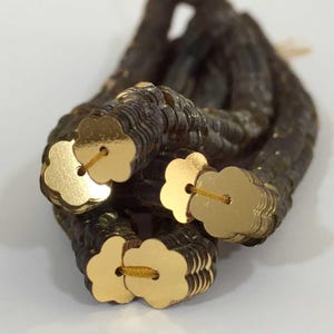 May include: A close-up of a strand of gold flower-shaped sequins. The sequins are strung together on a thin, dark brown thread.