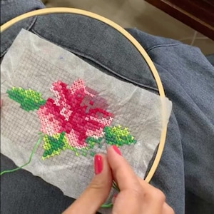Puede incluir: Primer plano de un proyecto de bordado de punto de cruz en progreso, con una rosa rosa con hojas verdes. El bordado se está trabajando en un aro de bordado de madera.
