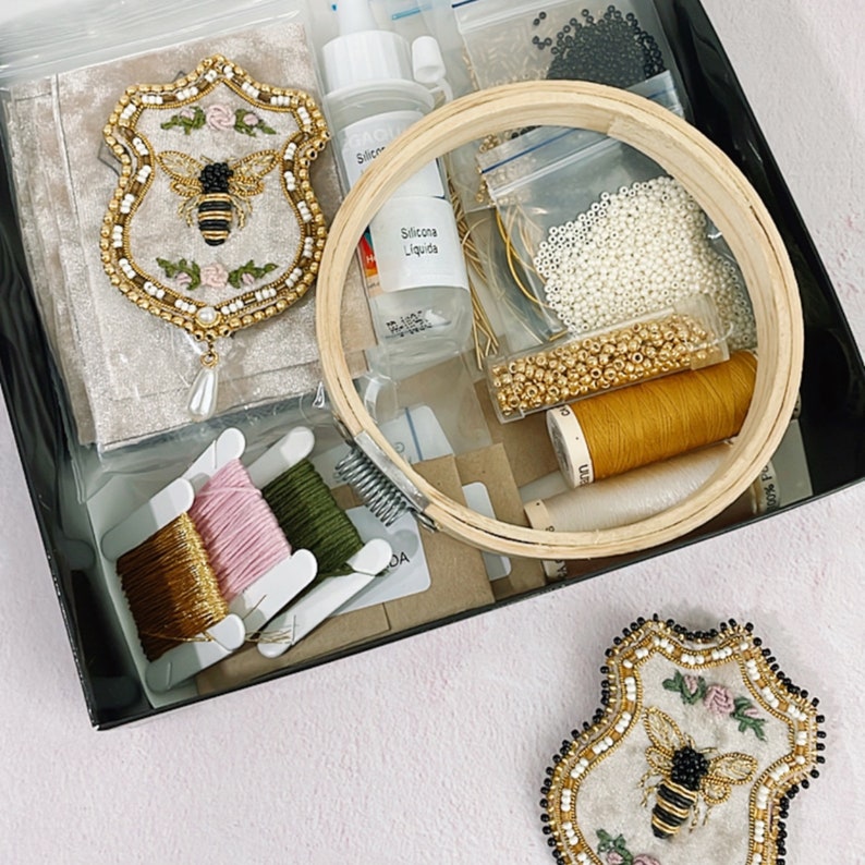 May include: A close-up of a craft kit for making embroidered bee brooches. The kit includes two pre-cut felt brooches, beads, thread, and a wooden embroidery hoop. The kit also includes a bottle of silicone liquid.