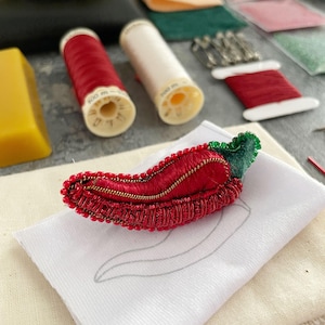 Può includere: Un broche a forma di peperoncino rosso e verde con perline, con un dettaglio di cerniera, è esposto su una superficie di tessuto bianco. Il broche è circondato da materiali per la cucitura, tra cui bobine di filo, aghi e un pezzo di tessuto.