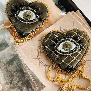 May include: Two green velvet heart-shaped brooches with gold chains and black beads. Each heart features an embroidered eye with a white iris, green pupil, and black lashes.