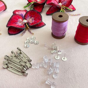 May include: A close-up of craft supplies for making jewelry or accessories. The image shows two spools of thread, one purple and one red, a pile of clear plastic earring backs, a pile of silver safety pins, a pile of silver earring posts, and a pile of clear rhinestones.