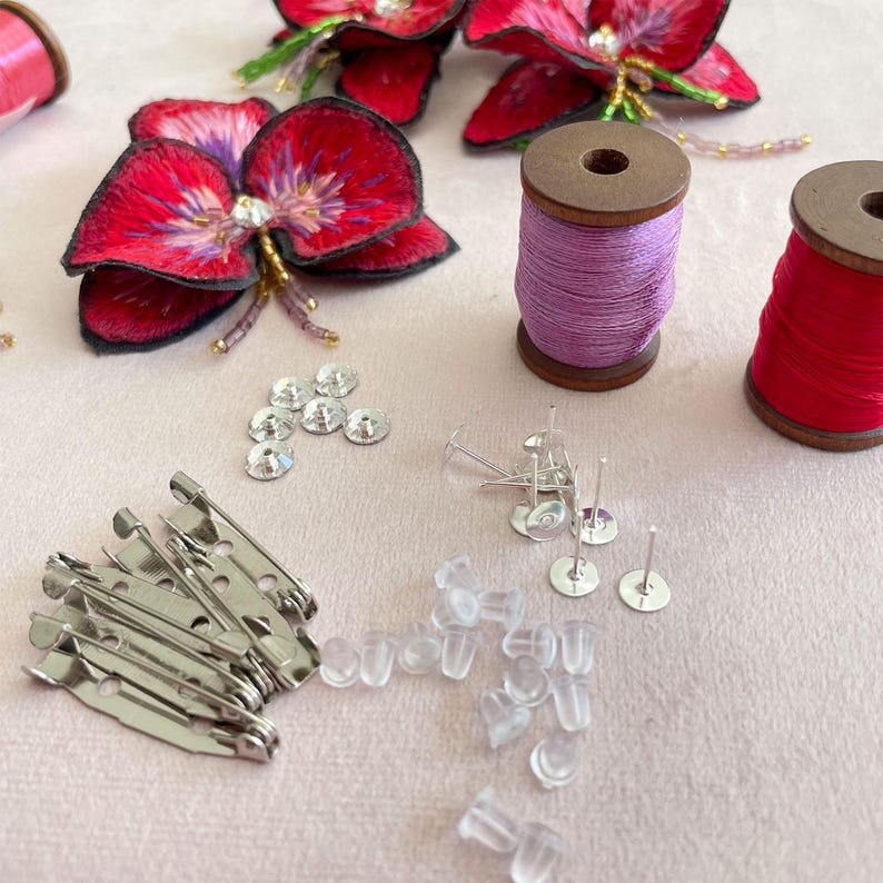 May include: A close-up of craft supplies for making jewelry or accessories. The image shows two spools of thread, one purple and one red, a pile of clear plastic earring backs, a pile of silver safety pins, a pile of silver earring posts, and a pile of clear rhinestones.