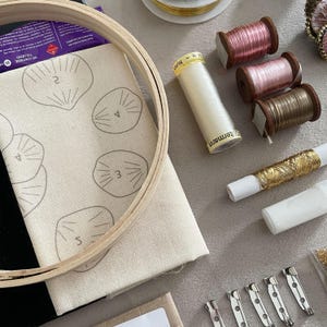 May include: A wooden embroidery hoop framing a piece of off-white fabric with printed numbered shell outlines, accompanied by spools of pink, brown, and gold thread, a white spool of G&uuml;termann nylon thread, and four silver metal brooch pin backs.