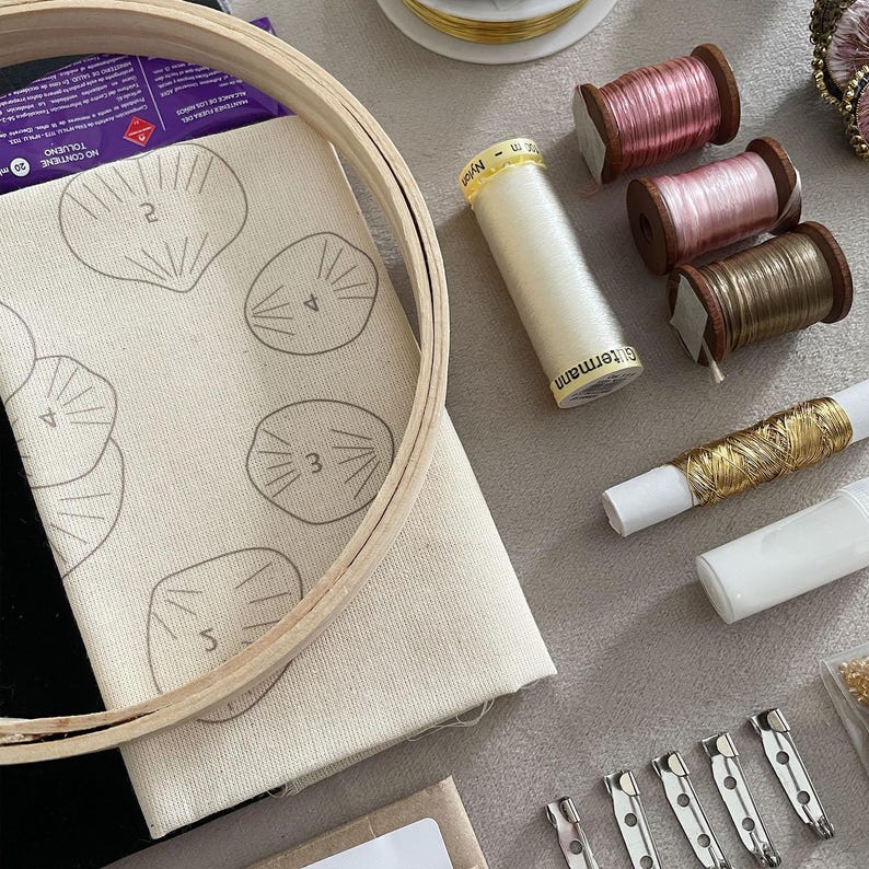 May include: A wooden embroidery hoop framing a piece of off-white fabric with printed numbered shell outlines, accompanied by spools of pink, brown, and gold thread, a white spool of G&uuml;termann nylon thread, and four silver metal brooch pin backs.