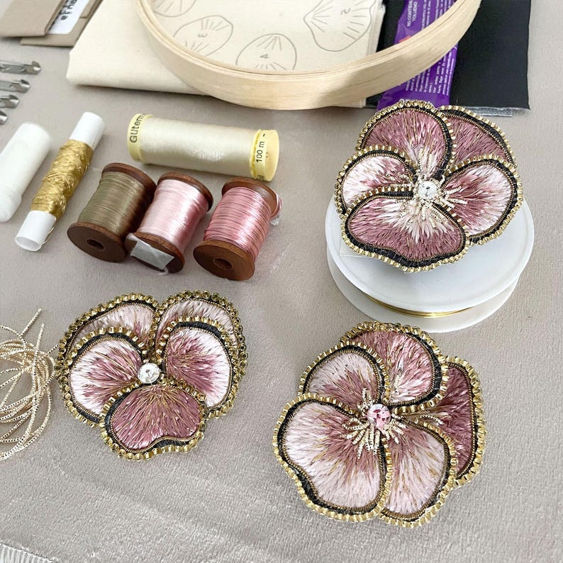 May include: Three embroidered pansy flower patches in shades of dusty pink and mauve with black stitched edges and a central crystal accent. The patches are displayed alongside various sewing supplies including spools of thread and an embroidery hoop.