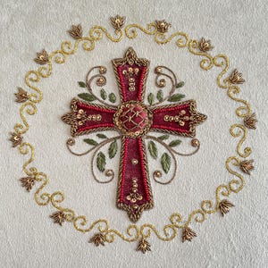 May include: Embroidered cross with a burgundy center, adorned with red beads and gold accents. The cross is surrounded by a gold floral design on a white background. The design includes green leaves.