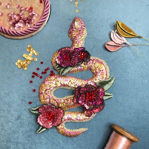 Puede incluir: Diseño de serpiente bordada con lentejuelas rosas, amarillas y doradas, adornada con flores rojas con lentejuelas y hojas verdes. La serpiente está sobre un fondo azul, con adornos adicionales.