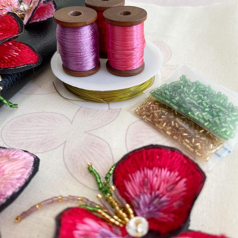 May include: Close-up of a craft project with two spools of pink and purple thread, a spool of gold wire, and two bags of green and gold beads. The beads are arranged in a clear plastic bag. The project is on a white fabric with a floral design.