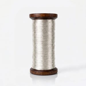 Puede incluir: Un carrete de hilo plateado metálico enrollado alrededor de una bobina de madera de color marrón oscuro. El hilo tiene una calidad reflectante y brillante, y está envuelto uniformemente alrededor del centro cilíndrico del carrete.