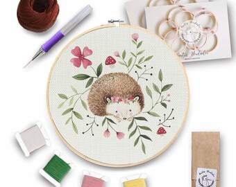 May include: A cross-stitch embroidery kit featuring a cute hedgehog wearing a flower crown and surrounded by greenery. The kit includes embroidery floss, a needle, a hoop, and instructions.