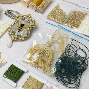 May include: A close-up of a bee embroidery kit. The kit includes a white felt bee with black wings, gold beads, green thread, gold thread, black thread, and gold wire.