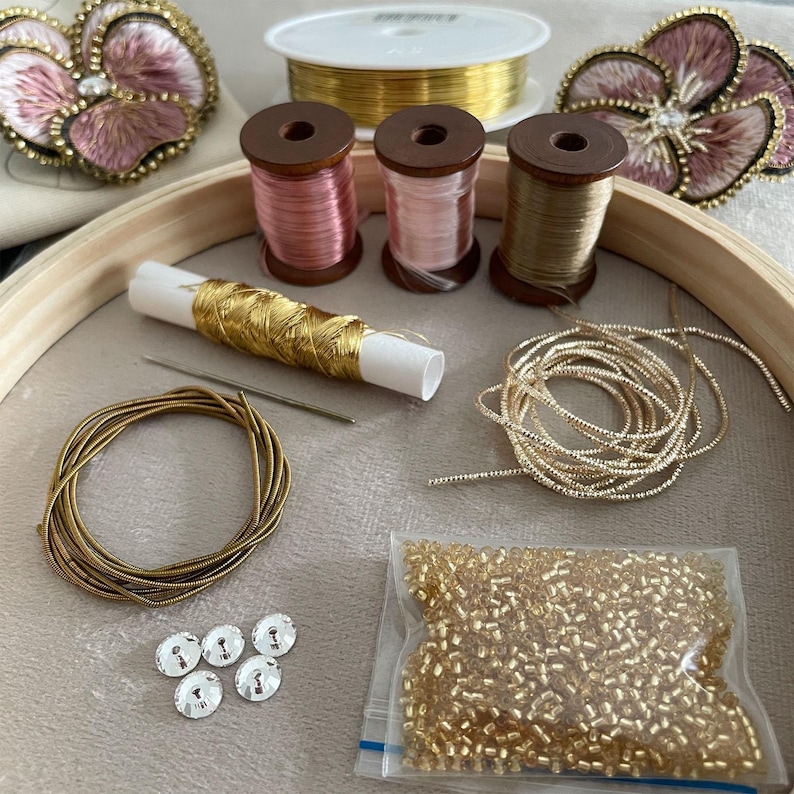 May include: An embroidery kit featuring two floral patches with pink thread and gold trim, three wooden spools of pink and tan thread, a spool of gold wire, a needle, gold metallic cord, a coil of gold gimp, five clear sequins, and gold beads.