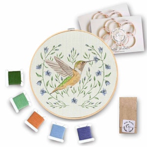 Puede incluir: Un kit de punto de cruz con un colibrí con plumas verdes y amarillas, rodeado de flores azules y hojas verdes. El kit incluye hilo de bordar en cinco colores, un aro de bordado de madera y una aguja.