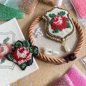 Puede incluir: Vista superior de broches florales con cuentas, incluido uno en forma de escudo con una rosa roja y otro diseño similar. Varias cuentas de colores, perlas y un ribete de cuerda rodean los broches, lo que sugiere materiales y suministros de artesanía.