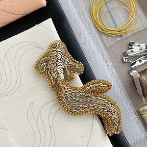 Puede incluir: Broche con forma de pez en oro y plata, con intrincados detalles de abalorios y hilos metálicos. La imagen muestra materiales de costura, como hilo, alfileres y tela.