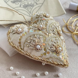 Puede incluir: Un adorno en forma de corazón, de color crema, con intrincados bordados dorados, lentejuelas y perlas de imitación. Los bordes están adornados con un cordón dorado. El adorno tiene una cuerda dorada para colgar.
