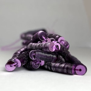 Puede incluir: Primer plano de un montón de lentejuelas moradas, cada una con un pequeño agujero para ensartar. Las lentejuelas son redondas y tienen un acabado brillante y metálico. El hilo es de color morado a juego.