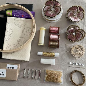 May include: An embroidery kit featuring a wooden hoop, fabric with printed shell outlines, three finished pansy-shaped fabric flowers, spools of thread in cream, pink, and brown, gold metallic thread, a needle, beads, and metal pin backings.