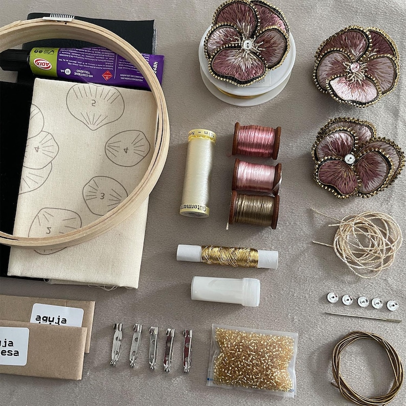 May include: An embroidery kit featuring a wooden hoop, fabric with printed shell outlines, three finished pansy-shaped fabric flowers, spools of thread in cream, pink, and brown, gold metallic thread, a needle, beads, and metal pin backings.