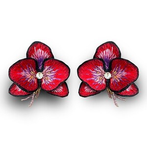 May include: A pair of red and purple embroidered flower earrings with a clear rhinestone center.