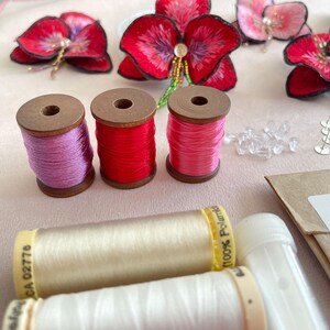 May include: Three spools of embroidery floss in pink, red, and light pink, and two spools of white embroidery floss. The spools are made of wood and have a label on the side.