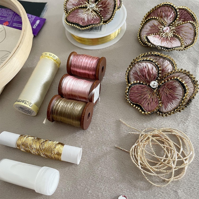 May include: Three embroidered pansy flower patches in shades of dusty pink and cream with gold beaded edges, alongside spools of pink, gold, and cream thread, a coil of gold metallic wire, and a wooden embroidery hoop on a light gray surface.