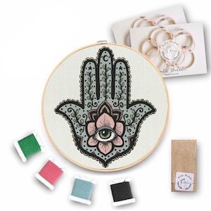 Puede incluir: Un kit de bordado pintado a mano con una mano de Hamsa con un diseño de ojo malvado. El kit incluye un aro, tela e hilo de bordar en tonos de verde, rosa y azul.