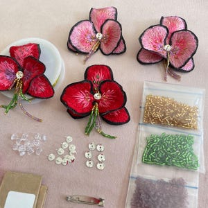 May include: Three pink and red embroidered flower brooches with rhinestones and beads. The brooches are surrounded by clear plastic push pins, silver sequins, and three bags of beads in gold, green, and purple.