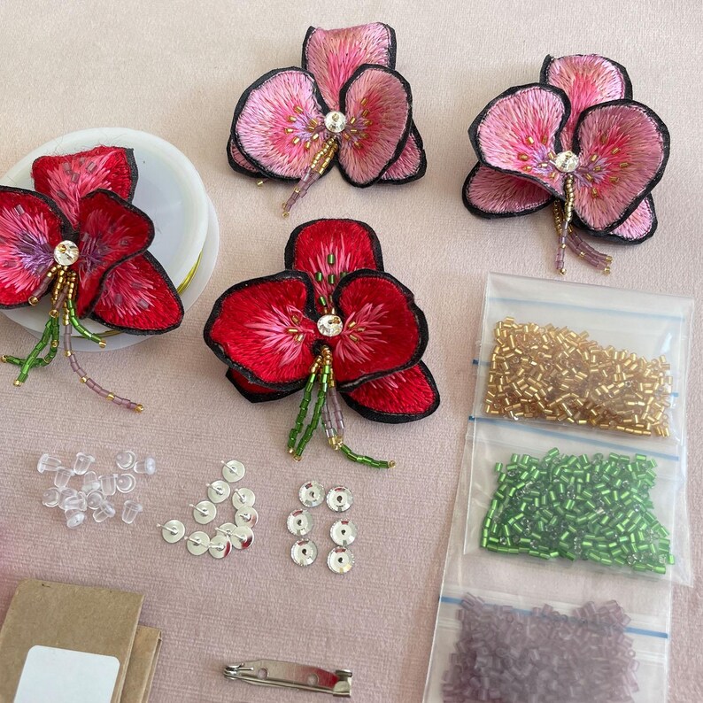 May include: Three pink and red embroidered flower brooches with rhinestones and beads. The brooches are surrounded by clear plastic push pins, silver sequins, and three bags of beads in gold, green, and purple.