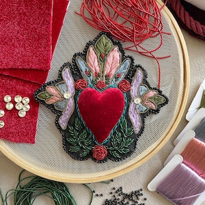 Puede incluir: Un corazón de terciopelo rojo con bordado de cuentas negras y hojas verdes, rodeado de plumas rosas, azules y verdes. El corazón está centrado en un aro de bordado de madera.