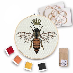 Puede incluir: Un kit de bordado de punto de cruz con una abeille con una corona en la cabeza. El kit incluye un aro de bordado de madera, tela Aida y cuatro madejas de hilo de bordar en rojo, naranja, amarillo y negro.