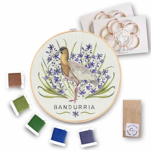 Puede incluir: Un kit de bordado de punto de cruz con una ilustración de acuarela de un pájaro Bandurria posado en un lecho de flores moradas. El kit incluye un aro de bordado de madera, tela Aida e hilo de bordar en marrón, verde, azul y morado.