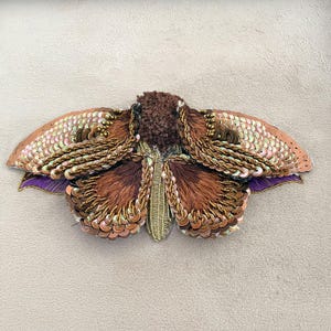 Puede incluir: Un parche bordado con forma de polilla que presenta un centro marrón difuso, lentejuelas de cobre iridiscentes y cuentas de oro metálico. Las alas están superpuestas con flecos de hilo marrón y detalles de tela morada oscura.