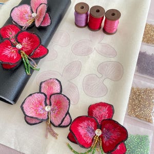 May include: Four embroidered orchid flower patches in pink and red with beaded accents. The patches are on a white fabric with printed orchid outlines. There are three spools of thread in pink, red, and white.