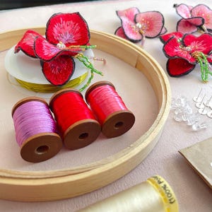 May include: A close-up of embroidery supplies including three spools of thread in pink, red, and purple, a wooden embroidery hoop, and several embroidered flower designs in pink and red.
