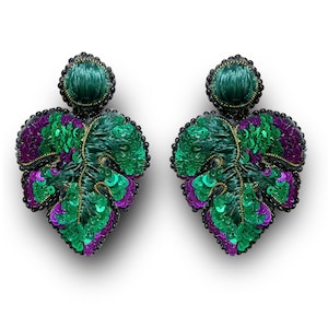 Puede incluir: Un par de pendientes con forma de hoja. Los pendientes presentan una combinación de lentejuelas verdes y moradas, con una parte superior verde oscuro y un borde de cuentas negras. Los pendientes están diseñados para parecerse a hojas.