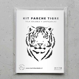Op de afbeelding: Een witte kaart met zwarte tekst die "Kit Parche Tigre Tela Soluble y Lentejuelas" zegt en een zwart-witte illustratie van een tijgergezicht. De kaart zit in een doorzichtige plastic zak.