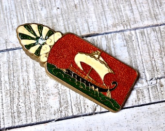 Puede incluir: Un marcapáginas de latón con un diseño de esmalte rojo, verde y blanco. El marcapáginas presenta un barco estilizado con una vela blanca y un casco verde y negro.
