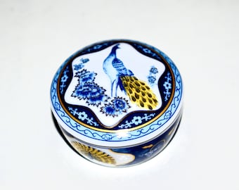 May include: A round, white porcelain trinket box with a blue and gold peacock design. The lid features a peacock with a spread tail, surrounded by blue and gold floral accents.