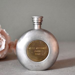 May include: A silver metal flask with a round body and a screw-on top. The flask has a brass circular plaque with the text "WEST WYCOMBE PARK 1998".